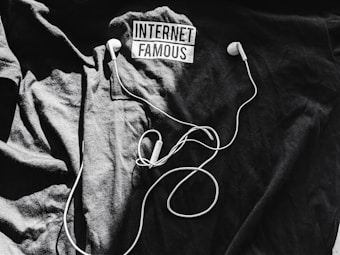 A black t-shirt is laid out with the words 'Internet Famous' printed on it. White earbuds with tangled cords rest on top of the shirt, creating a contrast against the dark background.
