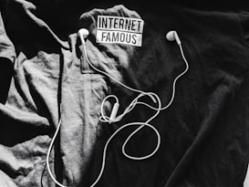 A black t-shirt is laid out with the words 'Internet Famous' printed on it. White earbuds with tangled cords rest on top of the shirt, creating a contrast against the dark background.