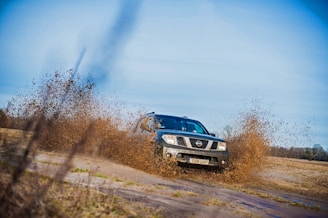 A vehicle is driving through a muddy field, splashing water and mud around it. The car, a Nissan, is positioned at an angle as it moves across the terrain, with muddy water spraying up around the wheels. The background consists of a vast open field and a blue sky.