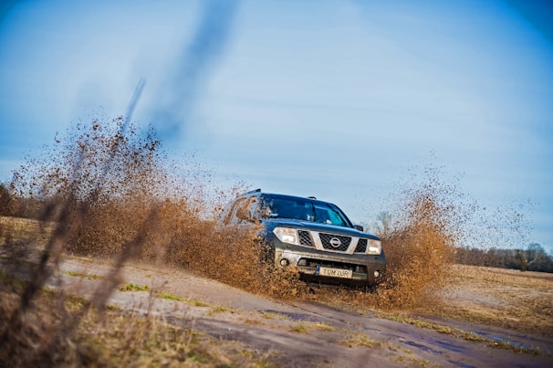 A vehicle is driving through a muddy field, splashing water and mud around it. The car, a Nissan, is positioned at an angle as it moves across the terrain, with muddy water spraying up around the wheels. The background consists of a vast open field and a blue sky.