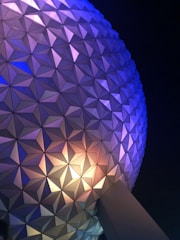A close-up view of a geodesic sphere structure with a triangular panel pattern illuminated with blue, purple, and yellow lights against a dark night sky.