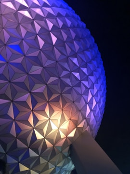 A close-up view of a geodesic sphere structure with a triangular panel pattern illuminated with blue, purple, and yellow lights against a dark night sky.