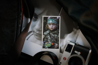 A close-up photograph of a printed image featuring a child in camouflage attire holding a toy gun, situated among other photographs. One noticeable item is a camera positioned at the bottom right corner.