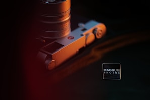 A vintage-style camera with a focus on its metallic details is partially visible, capturing the intricate design and the texture of its mechanical parts. The lighting is moody with a warm orange hue casting shadows and highlighting details. A small square badge with 'Magnum Photos' is placed nearby, suggesting a professional photography context.