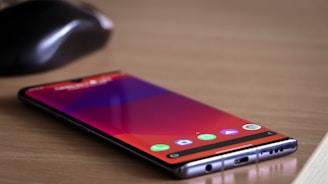 A smartphone lies flat on a wooden table with a screen displaying app icons. Near the phone, there is a black wireless computer mouse.