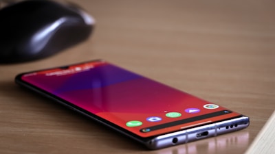 A smartphone lies flat on a wooden table with a screen displaying app icons. Near the phone, there is a black wireless computer mouse.