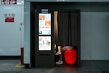 A photo booth in a public space with a bright display on one side showcasing a variety of advertisements and services, including passport and visa photo options. Inside the booth, partially visible legs of a person are seated on a glowing red stool with a curtain drawn partially closed. The booth is positioned against a white wall with fire safety signs nearby.