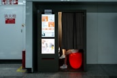 A photo booth in a public space with a bright display on one side showcasing a variety of advertisements and services, including passport and visa photo options. Inside the booth, partially visible legs of a person are seated on a glowing red stool with a curtain drawn partially closed. The booth is positioned against a white wall with fire safety signs nearby.