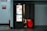 A photo booth in a public space with a bright display on one side showcasing a variety of advertisements and services, including passport and visa photo options. Inside the booth, partially visible legs of a person are seated on a glowing red stool with a curtain drawn partially closed. The booth is positioned against a white wall with fire safety signs nearby.
