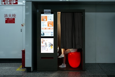 A photo booth in a public space with a bright display on one side showcasing a variety of advertisements and services, including passport and visa photo options. Inside the booth, partially visible legs of a person are seated on a glowing red stool with a curtain drawn partially closed. The booth is positioned against a white wall with fire safety signs nearby.