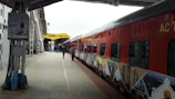 A red and grey train is stationary at a platform in a train station. Several passengers are seen walking on the platform, and the train features various scenic images painted along its side. A yellow sign labeled 'KAMAKHYA' is visible in the background, indicating the name of the station.