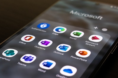 A smartphone screen displays a collection of Microsoft Office and associated applications, including icons for Outlook, Excel, Word, PowerPoint, OneNote, Microsoft Stream, SharePoint, and Teams. The apps are organized in a grid pattern with each icon being easily recognizable by its distinct color and design.