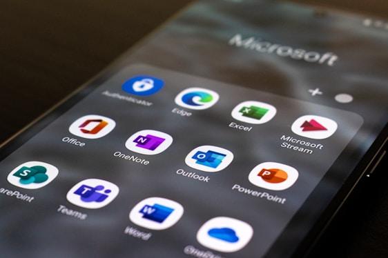 A smartphone screen displays a collection of Microsoft Office and associated applications, including icons for Outlook, Excel, Word, PowerPoint, OneNote, Microsoft Stream, SharePoint, and Teams. The apps are organized in a grid pattern with each icon being easily recognizable by its distinct color and design.