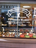 A patisserie shop window displays a variety of pastries and cakes, including colorful tarts and small, intricately decorated pastries. The signage on the window reads 'Artisans Pâtissiers' and 'Pâtisserie Maison,' indicating a focus on artisanal baked goods. Reflections of people and structures are visible in the glass.