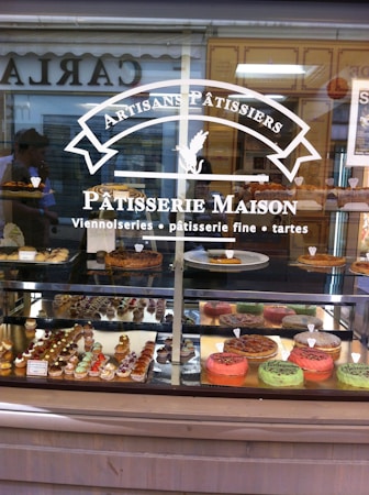 A patisserie shop window displays a variety of pastries and cakes, including colorful tarts and small, intricately decorated pastries. The signage on the window reads 'Artisans P&acirc;tissiers' and 'P&acirc;tisserie Maison,' indicating a focus on artisanal baked goods. Reflections of people and structures are visible in the glass.