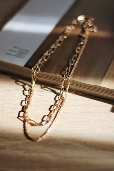 A delicate gold chain necklace lies on a wooden surface, casting subtle shadows. Sunlight illuminates the metal, creating a warm and inviting glow. A book rests beside the necklace, partially open, adding an element of casual elegance.