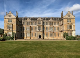 A large, historic mansion with an ornate facade featuring numerous windows and decorative stonework. The building has a symmetrical structure with two wings and a central section. It is surrounded by a well-manicured lawn and framed by a bright blue sky.