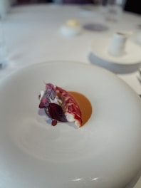A culinary dish features a delicately plated piece of lobster accompanied by artistic garnishes, including vibrant red leaves and a subtle sauce on a white plate. The setting is minimalist and elegant, suggesting a fine dining experience.