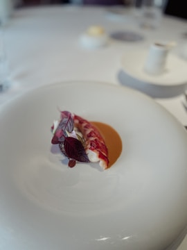 A culinary dish features a delicately plated piece of lobster accompanied by artistic garnishes, including vibrant red leaves and a subtle sauce on a white plate. The setting is minimalist and elegant, suggesting a fine dining experience.
