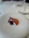 A culinary dish features a delicately plated piece of lobster accompanied by artistic garnishes, including vibrant red leaves and a subtle sauce on a white plate. The setting is minimalist and elegant, suggesting a fine dining experience.