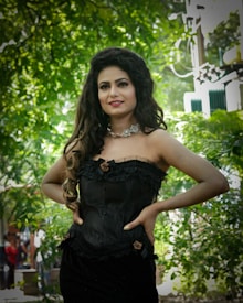 A woman stands confidently, wearing an elegant black corset dress adorned with floral details. Her long, wavy hair is styled beautifully, and she accessorizes with a delicate floral necklace. The background is lush with greenery, suggesting an outdoor setting, possibly a garden.