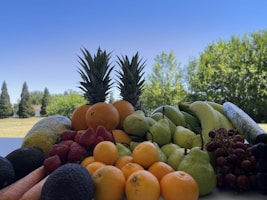 A variety of fresh fruits and vegetables are arranged on a table, including pineapples, oranges, strawberries, pears, bananas, grapes, avocados, carrots, and a cucumber. The background features a clear blue sky and lush green trees.