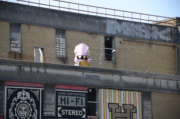 A brick building with industrial-style windows features a large toy figure resembling a dinosaur or monster emerging from the roof. Graffiti and murals decorate the building, including bold text and intricate designs.