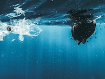 An underwater perspective reveals a clear view of the ocean surface with a variety of debris, including plastic waste, floating above. The water is illuminated by natural light, creating a serene yet unsettling view of pollution beneath the tranquility.