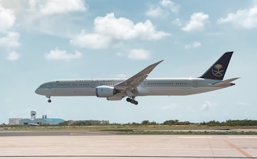 An aircraft from Saudia airlines is in mid-air, about to land at an airport. The background features a clear sky with scattered fluffy clouds. Below the plane, there is a runway and some airport buildings with a radar dome.