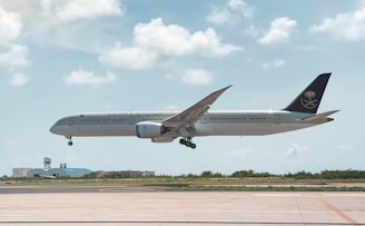 An aircraft from Saudia airlines is in mid-air, about to land at an airport. The background features a clear sky with scattered fluffy clouds. Below the plane, there is a runway and some airport buildings with a radar dome.