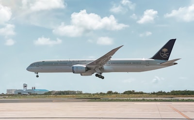 An aircraft from Saudia airlines is in mid-air, about to land at an airport. The background features a clear sky with scattered fluffy clouds. Below the plane, there is a runway and some airport buildings with a radar dome.