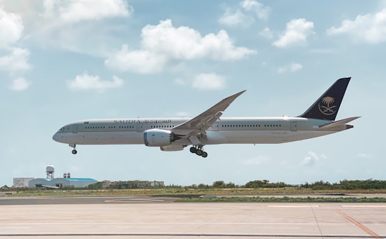 An aircraft from Saudia airlines is in mid-air, about to land at an airport. The background features a clear sky with scattered fluffy clouds. Below the plane, there is a runway and some airport buildings with a radar dome.