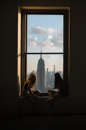 two girls sitting on a window sill looking out at a city