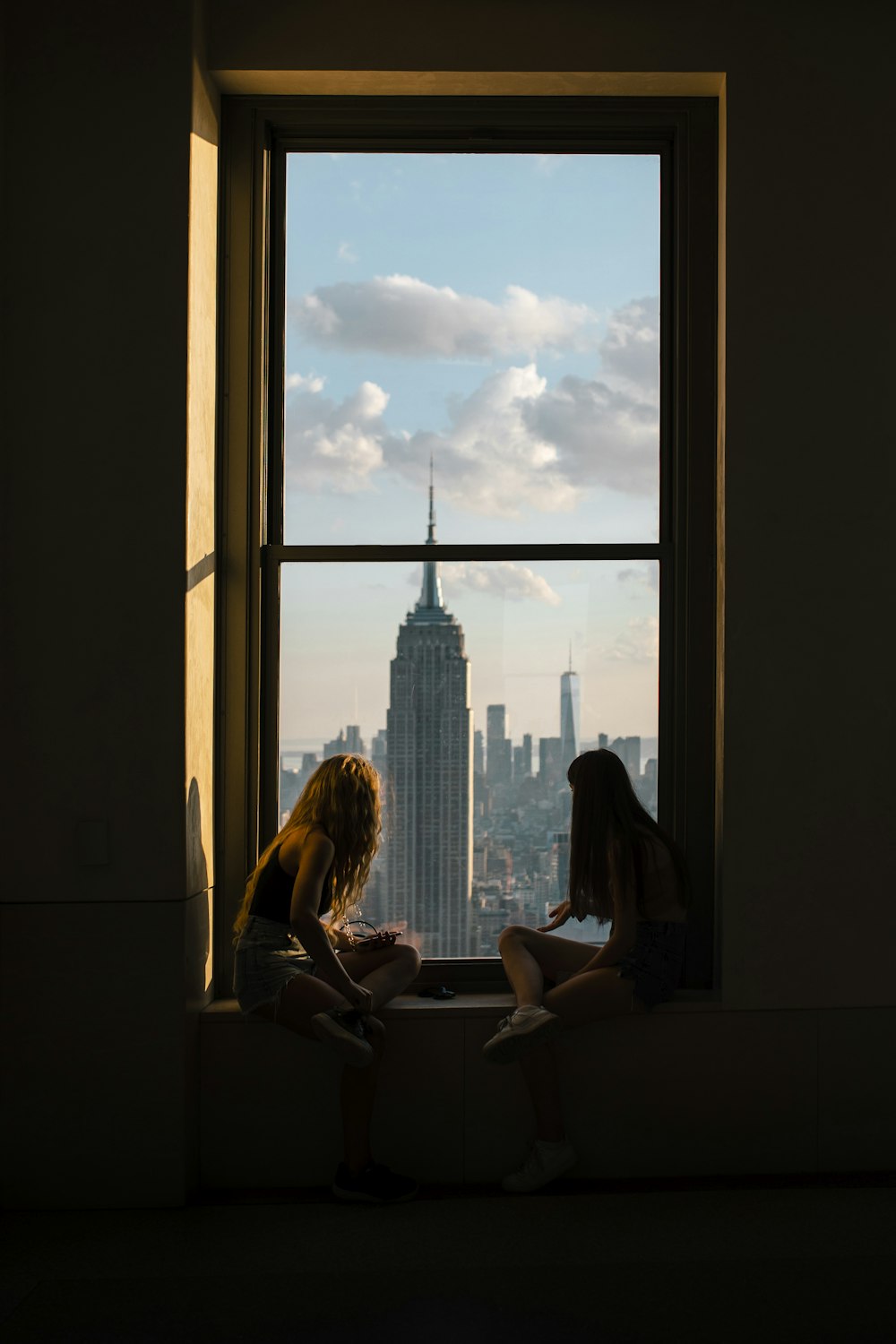 two girls sitting on a window sill looking out at a city