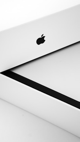 A close-up of a minimalistic white box with a clean design, featuring the iconic Apple logo prominently displayed in black. The box appears to be slightly open, revealing a sleek black interior.