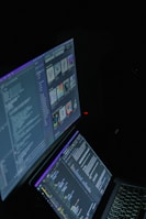 A dimly lit setting features two computer monitors displaying code and digital artwork. One monitor shows various programming code lines, while the other displays several digital images arranged in a grid-like format. Both screens are emitting a dark glow, suggesting a focused environment.
