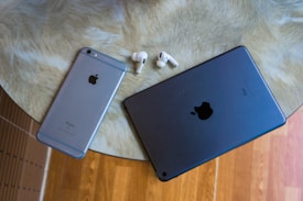 A round table with a beige textured surface holds an Apple iPhone, an iPad, and a pair of AirPods, showcasing a collection of tech gadgets. The background includes a wooden floor.