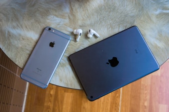 A round table with a beige textured surface holds an Apple iPhone, an iPad, and a pair of AirPods, showcasing a collection of tech gadgets. The background includes a wooden floor.