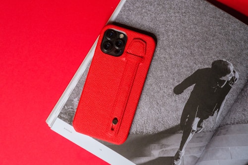 A red smartphone case placed on an open magazine with a black and white image. The image shows a person walking on steps, captured from a top-down perspective. The background is a strong red, with the magazine partially visible, providing contrast to the monochrome photo.