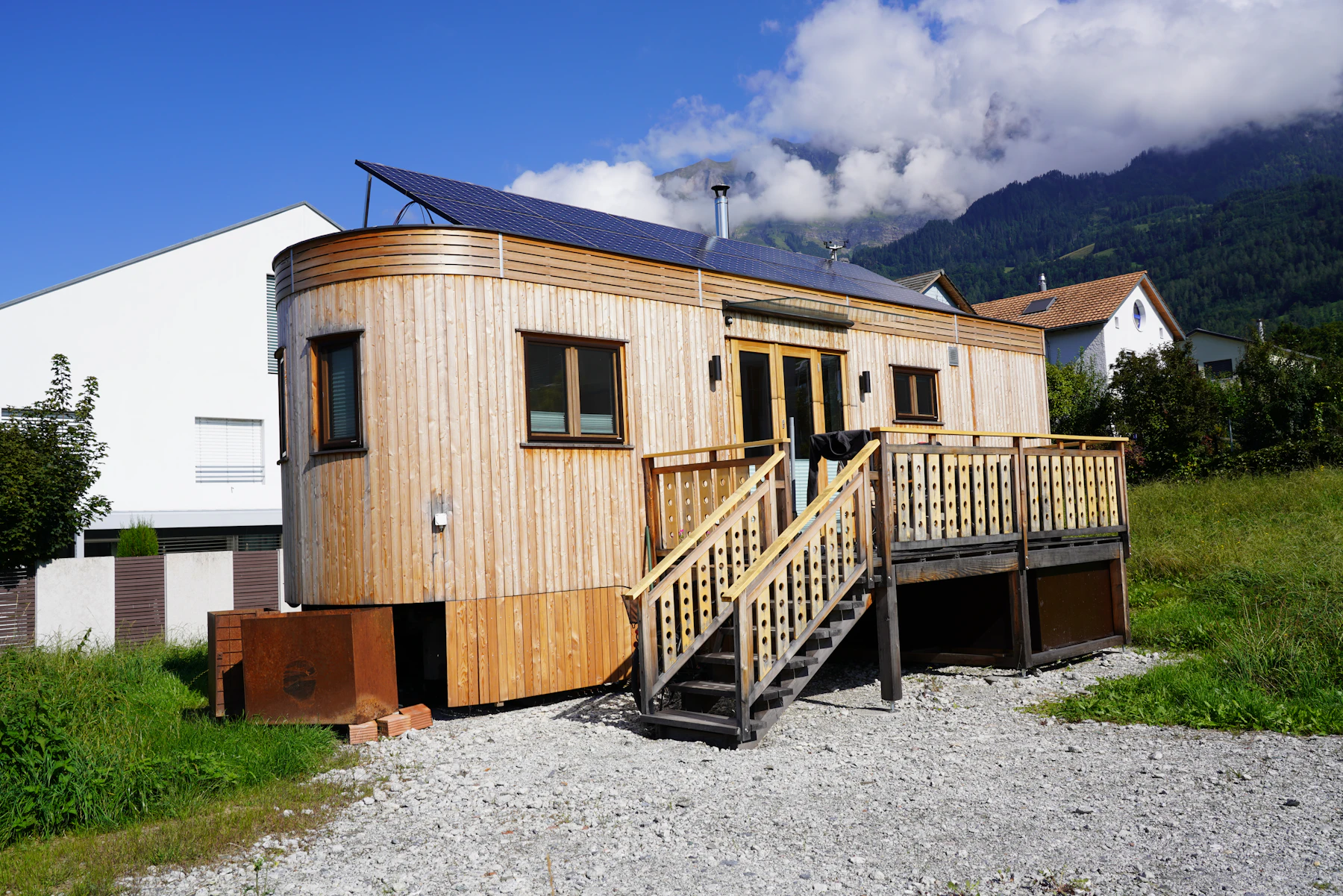 A wood-clad tiny house with rooftop solar panels and deck stairs, parked on gravel beside ordinary houses under mountains