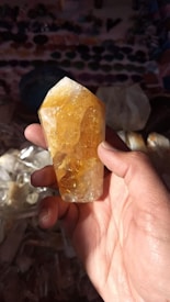 A hand holds a large, translucent crystal with a bright yellow hue. The crystal is faceted, catching the light and revealing internal textures. The background is filled with various other crystals and gemstones, slightly out of focus.