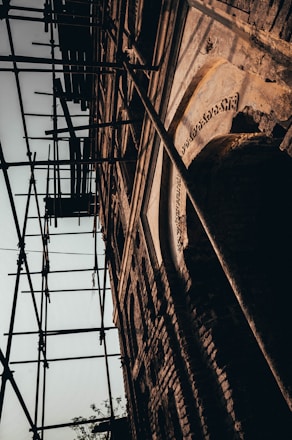 A close-up view of a brick building under renovation, with scaffolding around it. The structure displays intricate architectural details and weathered textures, suggesting age and historical significance.