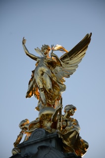 A golden statue of an angelic figure with large wings is perched atop a monument. The angel is holding a branch or laurel and is surrounded by additional figures, also in gold, beneath it. The background is a clear blue sky, highlighting the intricate details and shine of the gold surface.