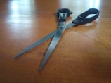 A pair of scissors with black handles rests on a wooden surface. The metal blades are slightly open, reflecting light from the surface. The texture and grain of the wood are visible, adding a rustic feel to the composition.