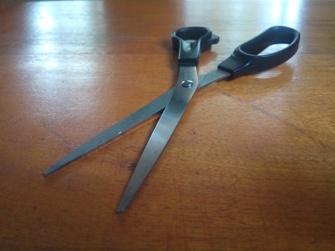 A pair of scissors with black handles rests on a wooden surface. The metal blades are slightly open, reflecting light from the surface. The texture and grain of the wood are visible, adding a rustic feel to the composition.