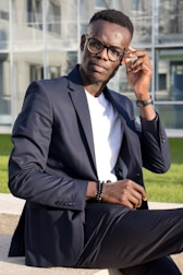 A person wearing a navy blue suit and glasses is sitting on a concrete bench outdoors. They are adjusting their glasses with one hand while looking towards the camera. The background features a modern glass building and a patch of green grass.