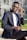 A person wearing a navy blue suit and glasses is sitting on a concrete bench outdoors. They are adjusting their glasses with one hand while looking towards the camera. The background features a modern glass building and a patch of green grass.