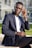 A person wearing a navy blue suit and glasses is sitting on a concrete bench outdoors. They are adjusting their glasses with one hand while looking towards the camera. The background features a modern glass building and a patch of green grass.