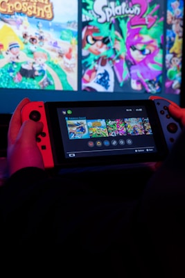 A person is holding a Nintendo Switch console, which displays a selection of video game titles on its screen. The console is shown with a red controller on the left and a blue controller on the right. The background features a larger screen displaying vibrant promotional graphics for games like Animal Crossing and Splatoon.