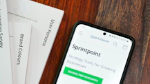 A wooden table with several documents and a smartphone. The documents are titled 'User Persona', 'Brand Colours', and 'Sprintpoint'. The smartphone displays a webpage for 'Sprintpoint', which offers strategy tools for growing businesses.